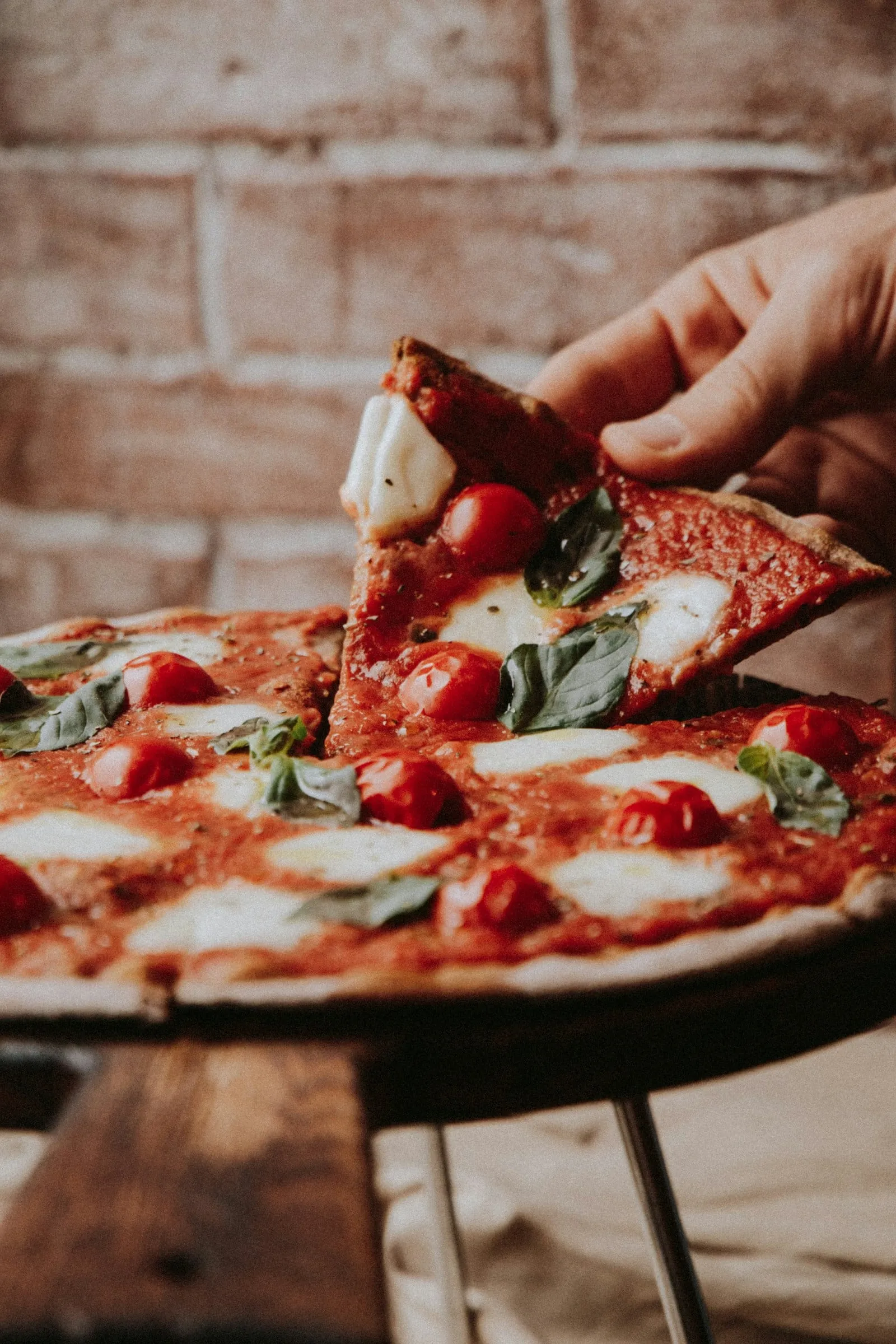 A hand lifting a slice of margherita in front of a brick wall