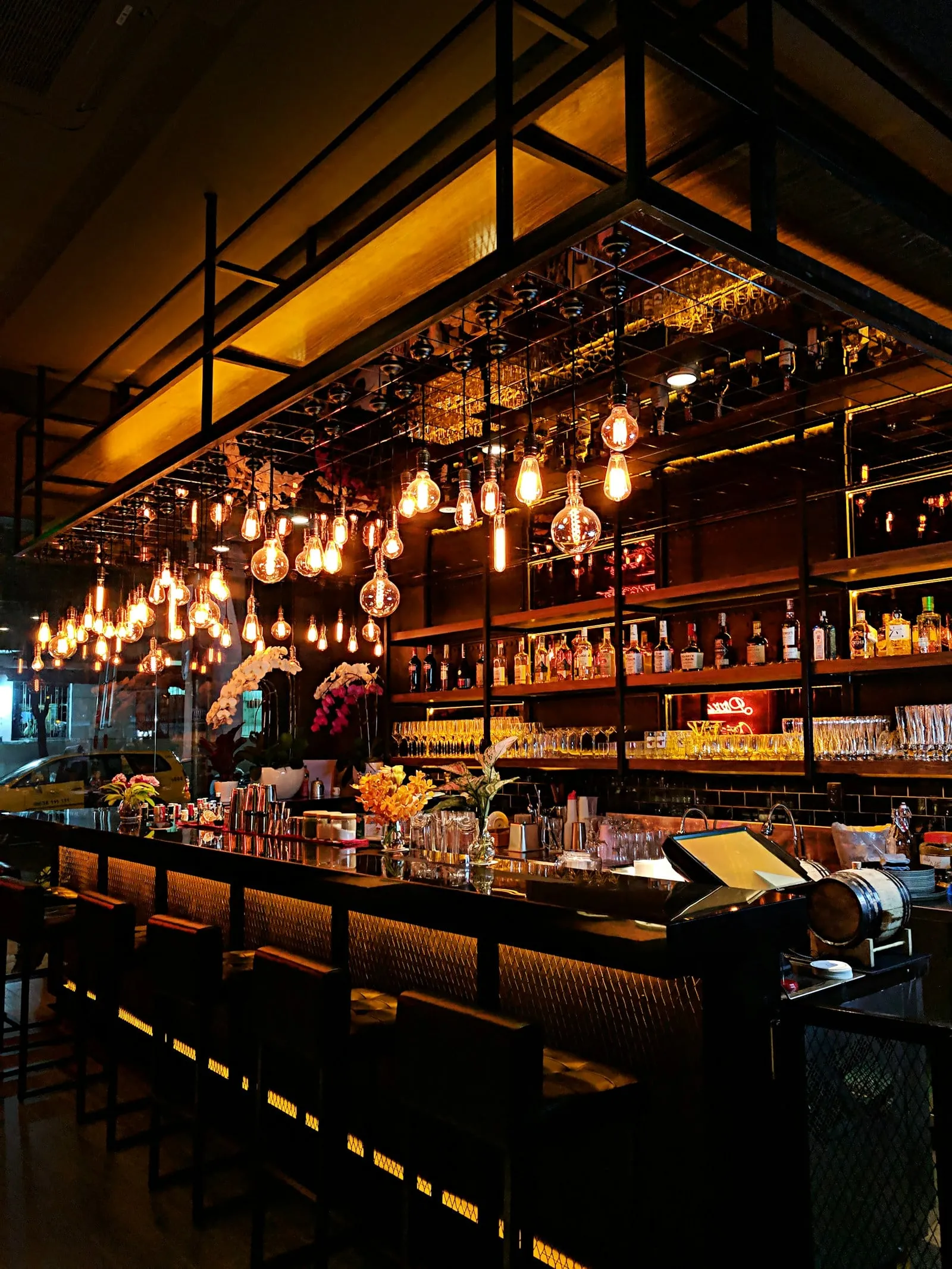 A long bar lit by hanging bulbs with shelves of bottles behind it